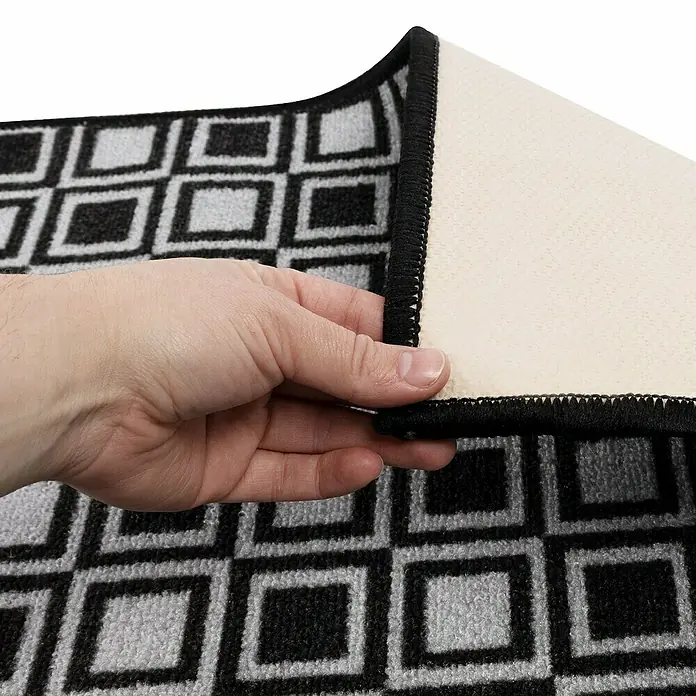 Teppichläufer Naas - Schwarz/Grau, 500 x 80 cm, 100 % Polyamid - Nahaufnahme einer Fußmatte mit geometrischem Muster, die von einer Hand angehoben wird, um die Rückseite zu zeigen.