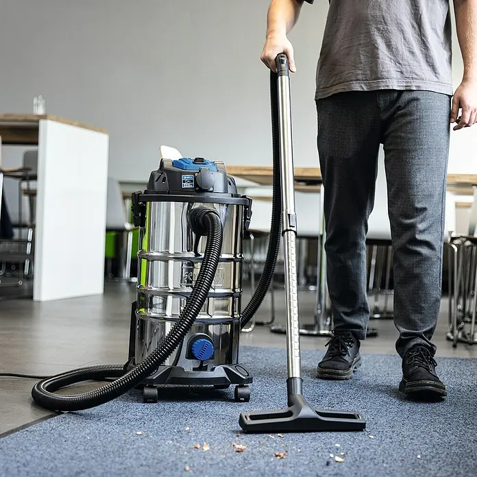 Scheppach Elektro-Nass- & Trockensauger VC30 Premium - 1.500 W, Behältervolumen: 30 l, Halbautomatisch - Ein Mann benutzt einen Nass- und Trockensauger mit Edelstahlbehälter auf einem Teppich. Der Sauger ist kompakt und leistungsstark.