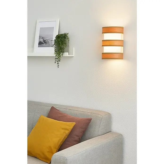 Tween Light Piano Aplique de pared - 40 W, An x Al: 15 x 20 cm, Beige, E27 - Una moderna lámpara de pared de madera con luz cálida acentúa un acogedor rincón de sala de estar con cojines y una planta en un estante.