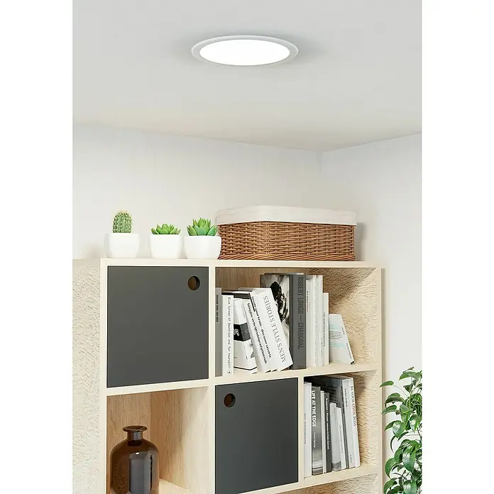 Eglo Fueva 6 Downlight empotrable LED redondo - 12 W, Ø x Al: 21,6 x 3,1 cm, Blanco, 2.700 K - 6.500 K, IP20 - Primer plano de una estantería con libros, decoraciones y una lámpara de techo redonda, que proyecta una luz brillante en la habitación.