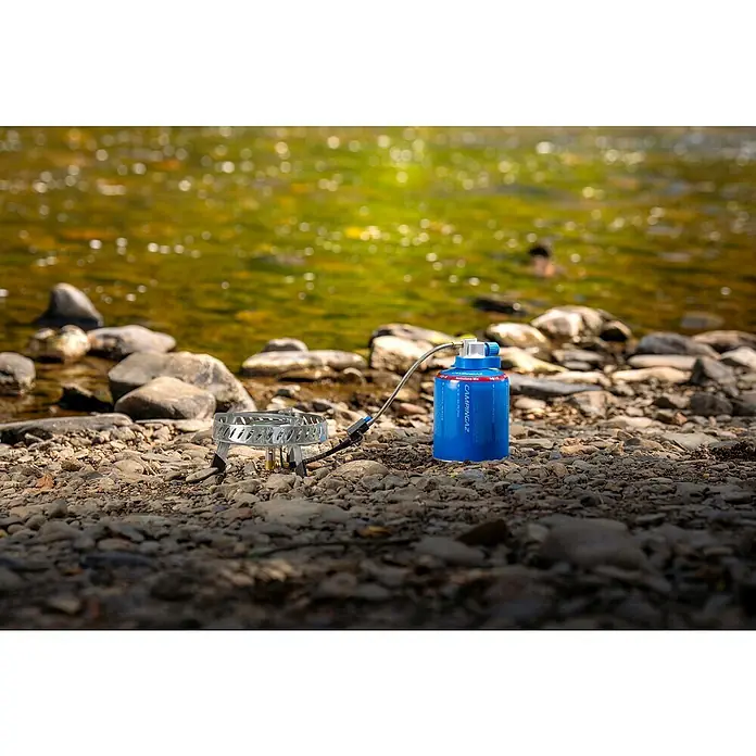 Campingaz Campingkocher Camp & Trek Connect CV - Anzahl Brenner: 1 Stk., 2 kW - Nahaufnahme eines Campingaz-Gaskochers mit blauer Gaskartusche am steinigen Ufer, ideal für Outdoor-Abenteuer und Camping.