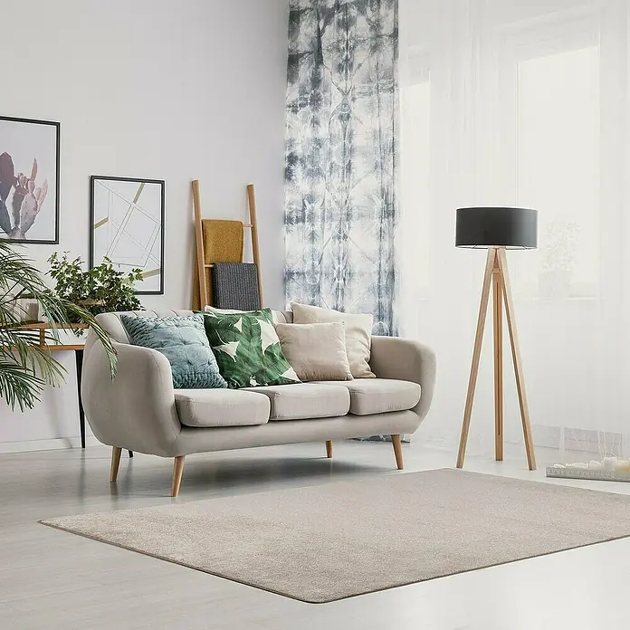 Kurzflorteppich Sundae - Beige, 200 x 200 cm, 100 % Polypropylen - Ein modernes Wohnzimmer mit einem beigen Sofa, Kissen und einem hellen Teppich. Eine Stehlampe mit Holzbeinen steht neben dem Fenster.