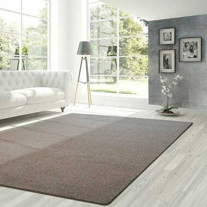 Kurzflorteppich Lyon - Braun, 450 x 80 cm, 100 % Polypropylen - Teppich in Braun-Grau in einem hellen Wohnzimmer mit Sofa und großen Fenstern. Der Teppich liegt auf einem hellen Holzboden.
