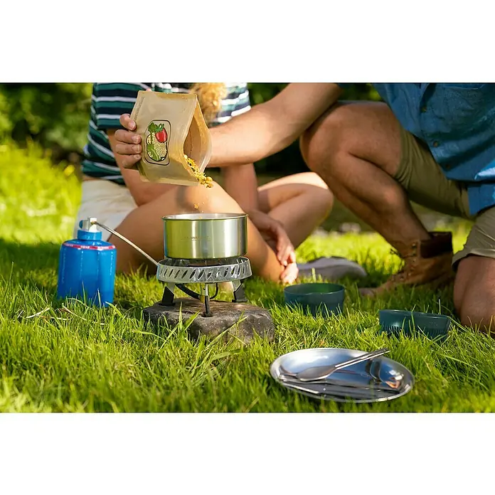 Campingaz Campingkocher Camp & Trek Connect CV - Anzahl Brenner: 1 Stk., 2 kW - Zwei Personen bereiten eine Mahlzeit mit einem Campingkocher und einem Lebensmittelsack im Freien zu. Campinggeschirr liegt im Gras bereit.