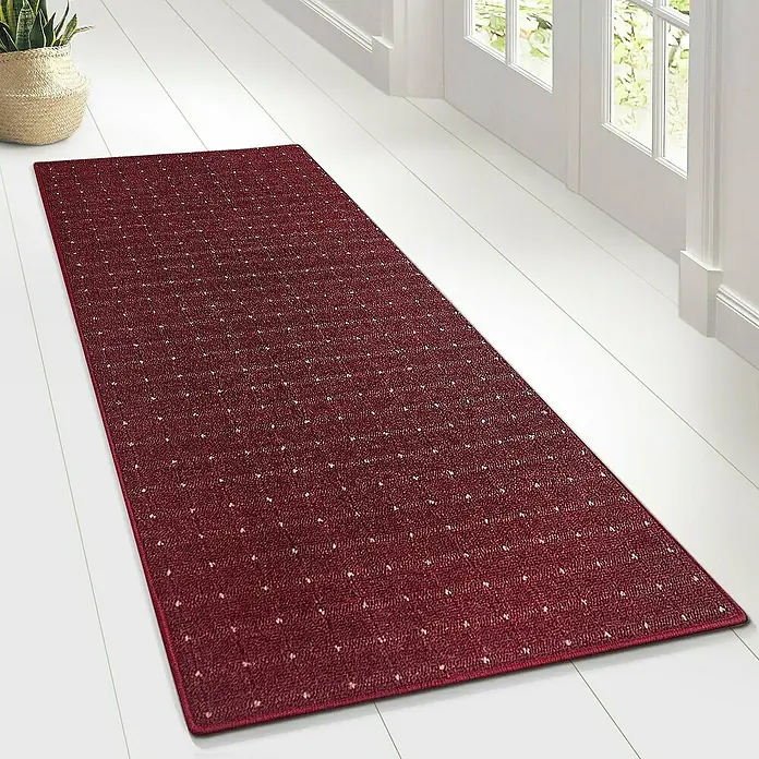 Flachgewebeteppich Rapido - Violett, 200 x 200 cm, 100 % Polypropylen - Ein roter Läufer mit dezentem Punktmuster liegt auf einem hellen Holzboden, neben einer Tür und einem Korb mit einer Grünpflanze.