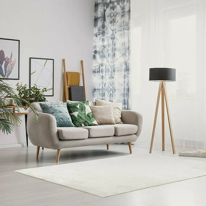 Kurzflorteppich Sundae - Creme, 200 x 200 cm, 100 % Polypropylen - Ein gemütliches Wohnzimmer mit einem hellen Teppich, einem beigen Sofa und einer Stehlampe, die für eine entspannte Atmosphäre sorgen.
