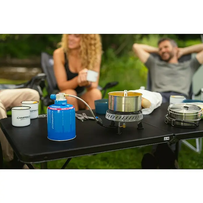 Campingaz Campingkocher Camp & Trek Connect CV - Anzahl Brenner: 1 Stk., 2 kW - Campingkocher mit Topf und Gaskartusche auf einem Tisch, umgeben von entspannten Personen im Hintergrund.