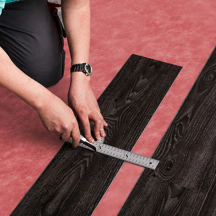 b!design Unterlagsmatte - 10 m², Stärke: 1,4 mm - Mann schneidet dunkle Vinyl-Bodenfliese mit einem Teppichmesser und einem Lineal.