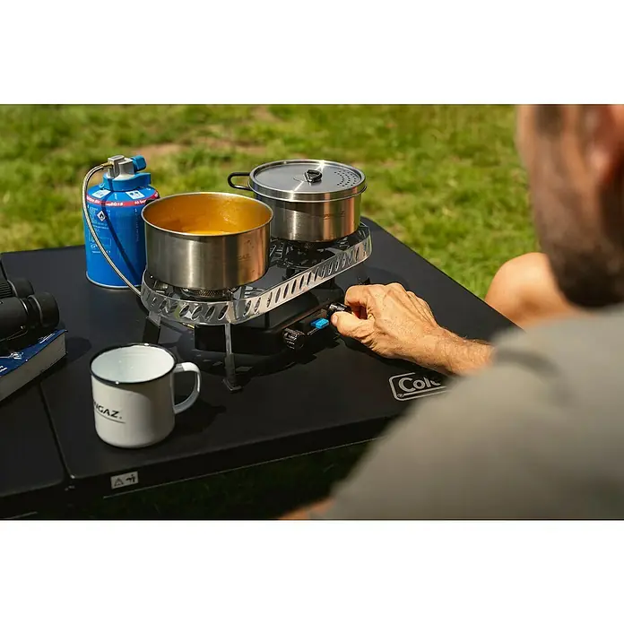 Campingaz Campingkocher Camp & Trek Twin Connect CV - Anzahl Brenner: 2 Stk., 2 kW - Eine Nahaufnahme eines Coleman-Campingkochers mit zwei Töpfen, der auf einem schwarzen Tisch im Freien steht, mit einer Hand, die einen Knopf bedient.