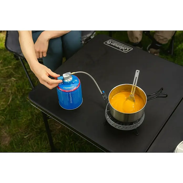 Campingaz Campingkocher Camp & Trek Connect CV - Anzahl Brenner: 1 Stk., 2 kW - Eine Person kocht mit einem Campingkocher auf einem schwarzen Coleman Tisch im Freien. Der Fokus liegt auf dem blauen Campingkocher.
