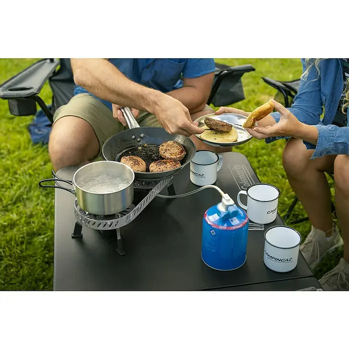 Campingaz Campingkocher Camp & Trek Twin Connect CV - Anzahl Brenner: 2 Stk., 2 kW - Ein tragbarer Campingkocher der Marke Coleman wird zum Kochen von Burgern auf einem Tisch im Freien verwendet.