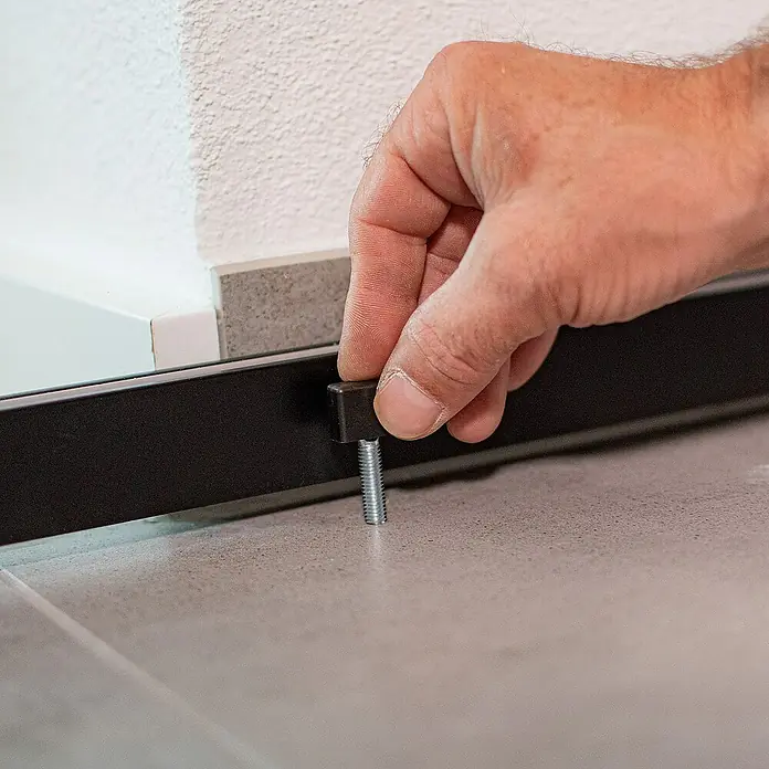 Inova Schiebetür-Komplettset Glasschiebetür ESG 755 x 2035 mm Schwarz gerahmt mit Softclose - 755 x 2.035 mm - Hand montiert einen schwarzen Nivellierfuß mit Schraube an einem Möbelstück auf einem grauen Boden.