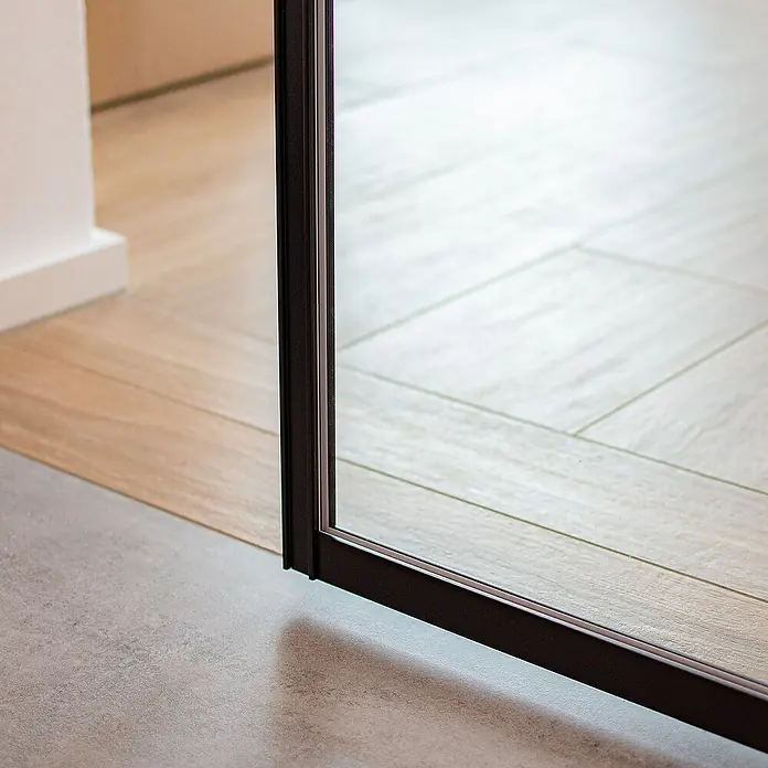 Inova Schiebetür-Komplettset Glasschiebetür ESG 880 x 2035 mm 3 Felder - 880 x 2.035 mm - Nahaufnahme einer Schiebetür aus dunklem Metall mit Glasfüllung, die zwischen Holzboden und grauem Fliesenboden abschließt.