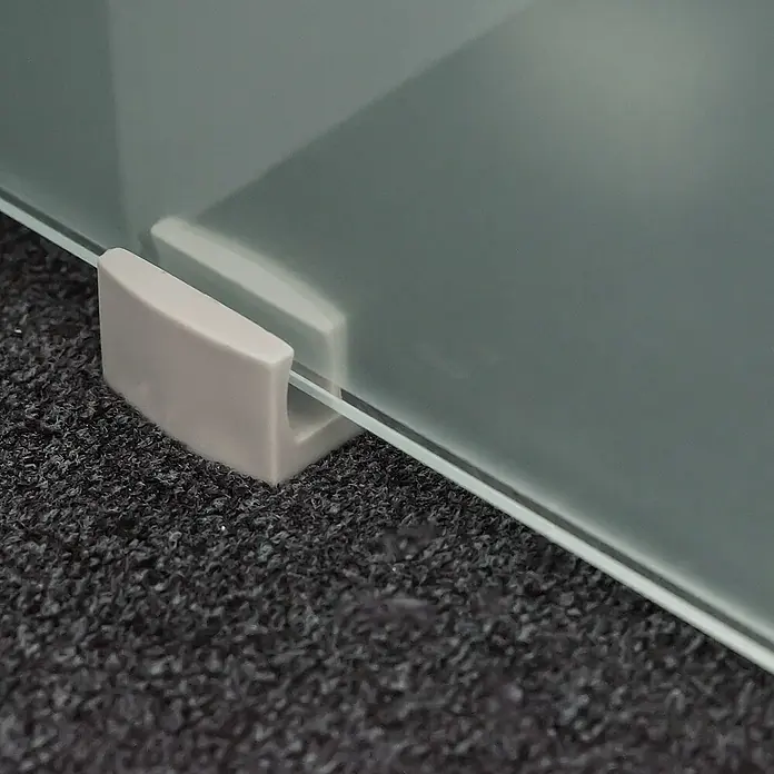 Inova Schiebetür-Komplettset Glas Komplettset wellenförmig mit Quadratgriff - 880 x 2.035 mm - Detailaufnahme einer Glasplatte, die mit einer hellgrauen Halterung auf einem dunklen, texturierten Untergrund befestigt ist.