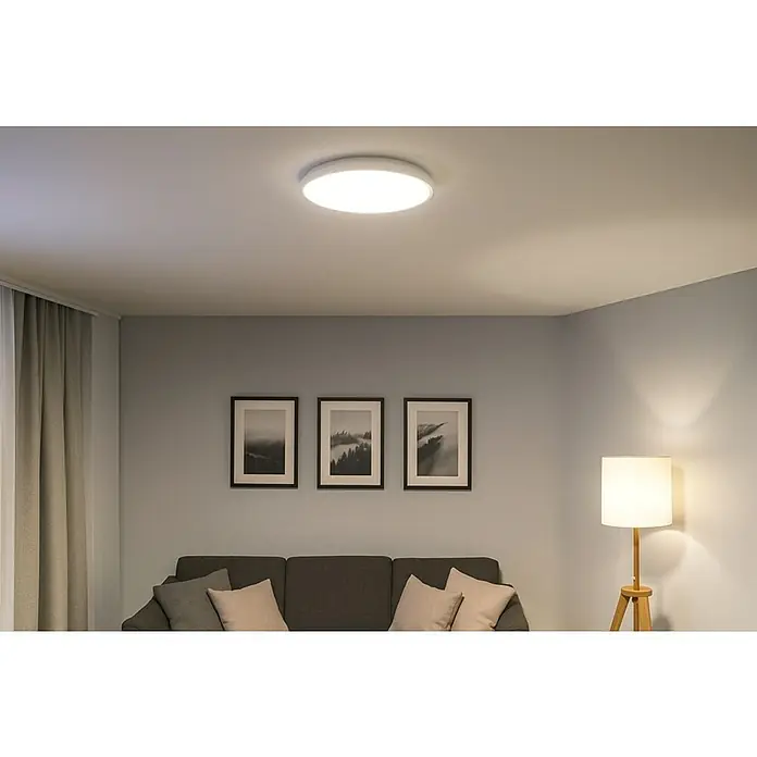 Eglo Plafón LED Testara-E - 11,2 W, Ø x Al: 21,1 x 3 cm, Blanco, Blanco neutro - Una lámpara de techo plana y circular brilla intensamente en una sala de estar decorada de forma moderna con un sofá gris y decoración minimalista.