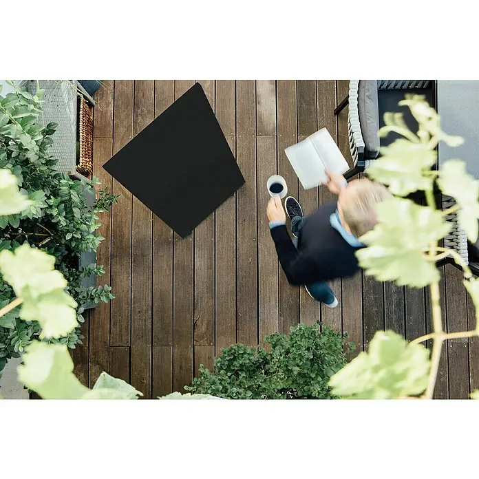 Jokerarea Infrarotheizkörper Area Warm 900 - L x B x H: 120 x 60 x 2,7 cm, 900 W, Schwarz - Draufsicht auf eine Terrasse mit einem Mann, der Kaffee trinkt und ein Buch liest, umgeben von Pflanzen und einem Tisch.
