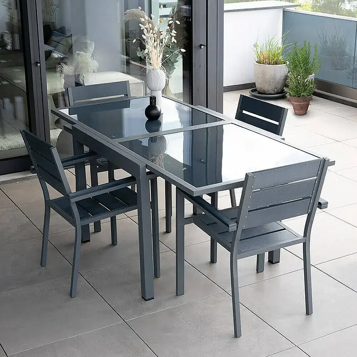 Home Deluxe Gartentisch-Set VERANO MADERA - Tisch inkl. 4 Stühlen - Gesamtumfang: 5 -tlg., Ausziehbar - Ein graues Gartentischset mit Glasplatte und passenden Stühlen auf einer Terrasse. Perfekt für Mahlzeiten im Freien.