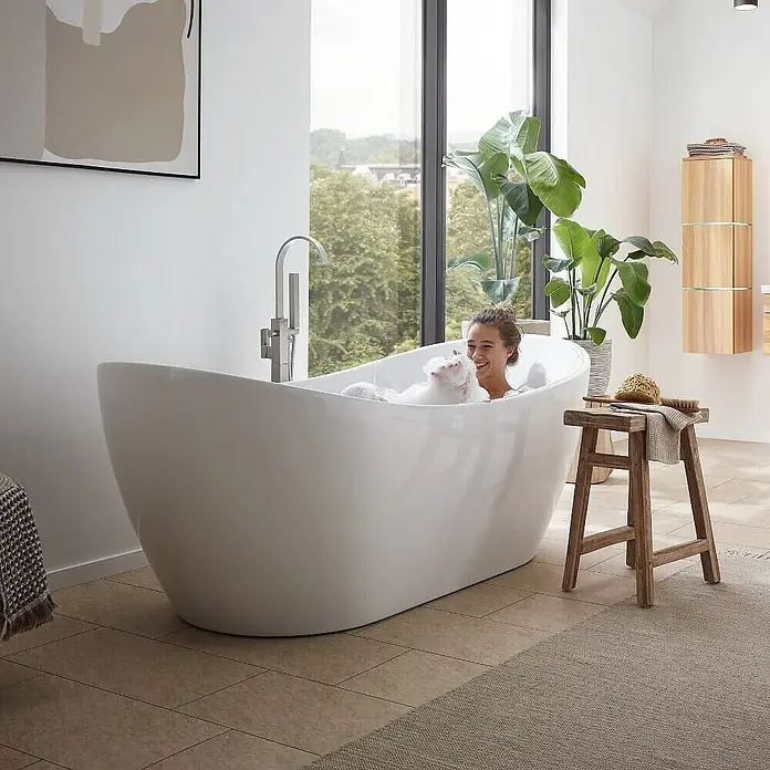 Home Deluxe Freistehende Badewanne OVALO PLUS - Weiß - Frau entspannt in einer freistehenden Badewanne mit Schaum. Das Badezimmer verfügt über große Fenster und moderne Armaturen.