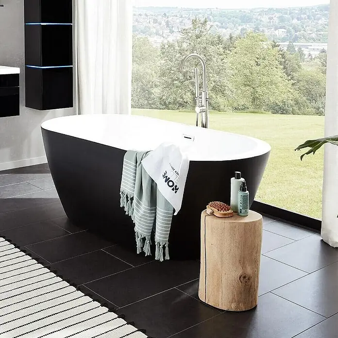 Home Deluxe Freistehende Badewanne CODO - Schwarz - Freistehende Badewanne in Schwarz und Weiß mit Handtuch und Holzschemel im Badezimmer mit Blick auf die Natur.