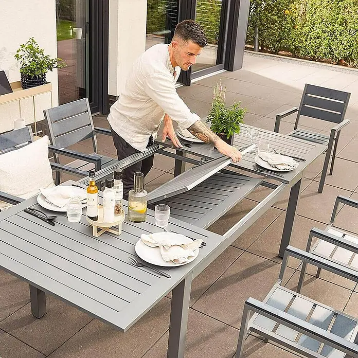 Home Deluxe Gartentisch-Set CASA MADERA - Tisch inkl. 4 Stühlen - Gesamtumfang: 5 -tlg., Ausziehbar - Ein Mann, der einen grauen Ausziehtisch auf einer Terrasse aufstellt. Der Tisch ist mit Geschirr gedeckt und von Stühlen umgeben.