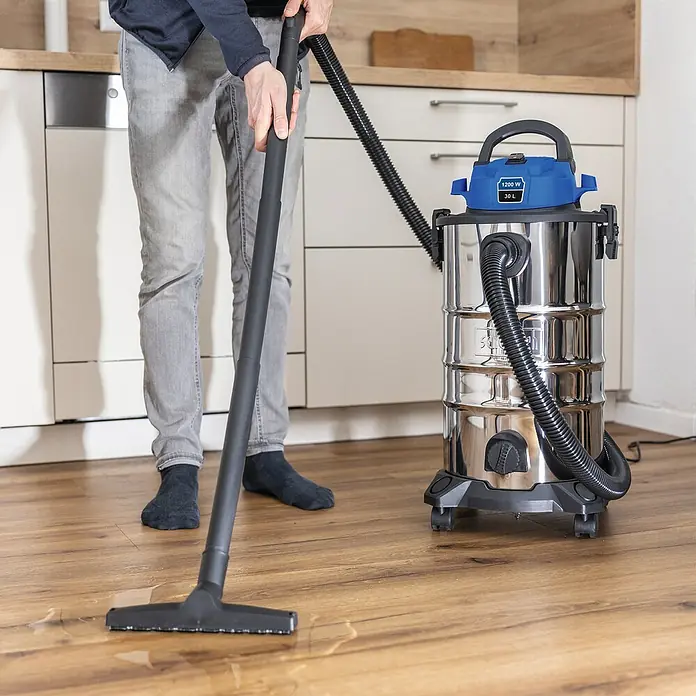 Scheppach Elektro-Nass- & Trockensauger VC30v2 - 1.200 W, Behältervolumen: 30 l, Manuell - Ein Mann benutzt einen Nass-Trockensauger zur Reinigung von Wasser auf einem Holzboden. Der Staubsauger ist aus Edelstahl mit blauem Oberteil.
