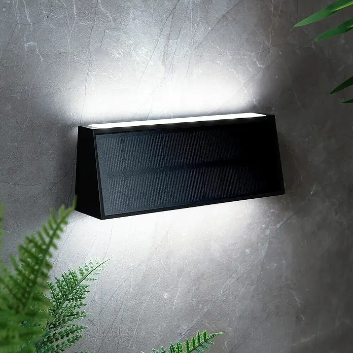 Arte confort Aplique solar para exterior Trape - 1,3 W, Blanco neutro, Negro - Lámpara de pared solar negra montada en una pared gris con plantas. Proporciona luz brillante hacia arriba y hacia abajo.