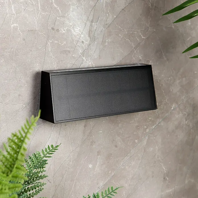 Arte confort Aplique solar para exterior Trape - 1,3 W, Blanco neutro, Negro - Una moderna lámpara solar negra para exteriores, montada en una pared gris y rodeada de plantas.