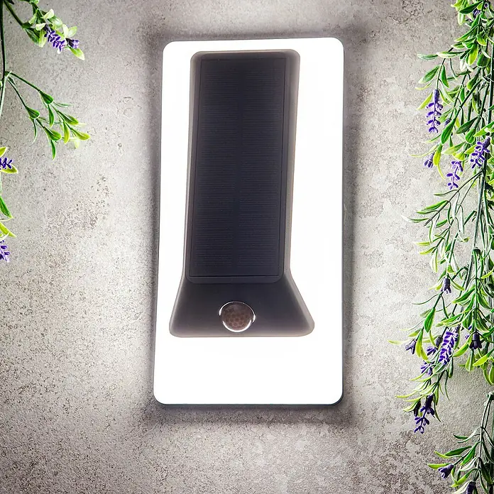 Arte confort Aplique solar para exterior Lexor - 1,4 W, Blanco cálido, Blanco - Primer plano de una lámpara de pared rectangular de energía solar con sensor de movimiento, rodeada de plantas decorativas.