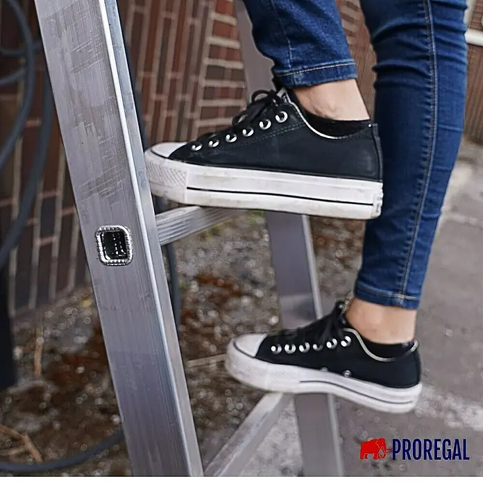 Proregal Anlegeleiter ATLAS - Anzahl Tritte: 9 Stk., Arbeitshöhe: 3.470 mm, Belastbarkeit: 150 kg, Aluminium - Eine Person, die auf einer silbernen PROREGAL-Leiter mit schwarzen Schuhen und Jeans steht.