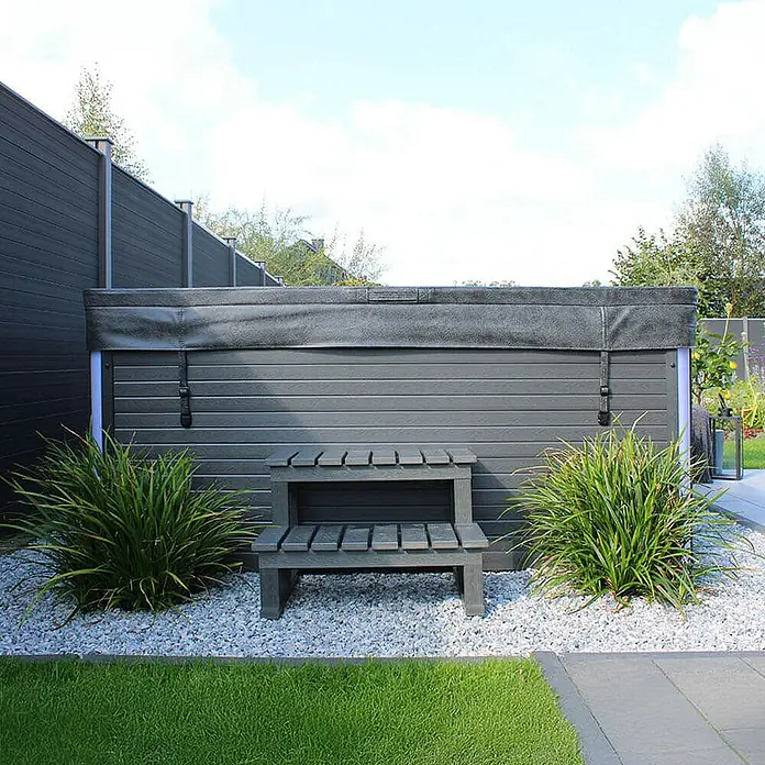 Home Deluxe Außen-Whirlpool STREAM BIG inkl. Treppe und Thermoabdeckung - 207,5 x 207,5 x 88 cm, Anthrazit, Max. Personenzahl: 5 Stk. - Diese Aufnahme zeigt einen Whirlpool mit Abdeckung, umgeben von dekorativen Pflanzen und einem kleinen Podest.