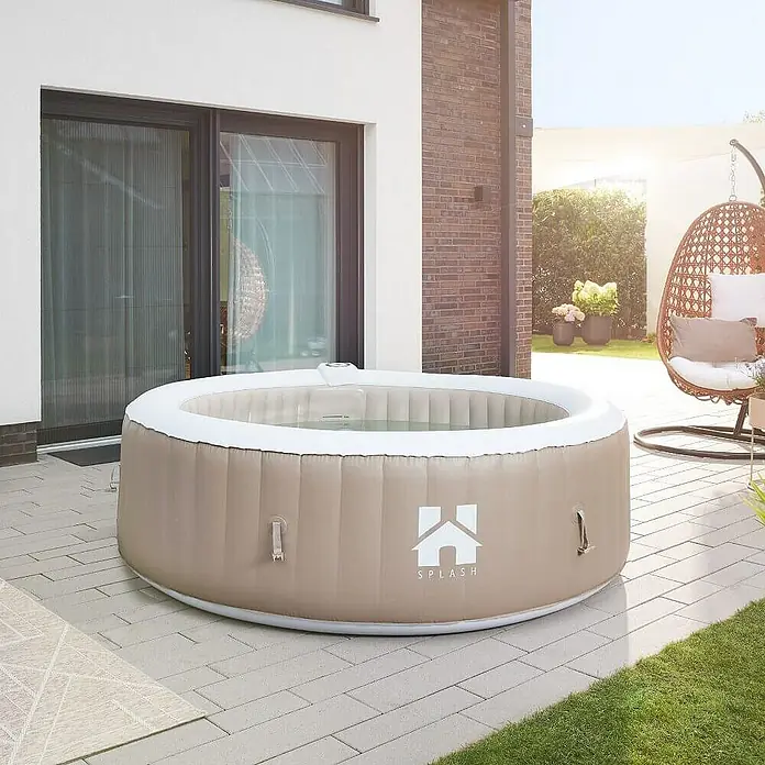 Home Deluxe Außen-Whirlpool SPLASH inkl. Abdeckung - 208 x 208 x 65 cm, Beige, Max. Personenzahl: 6 Stk. - Außenaufnahme eines beigen "Splash"-Whirlpools mit weißem Rand auf einer Terrasse mit einer Schaukel im Hintergrund.