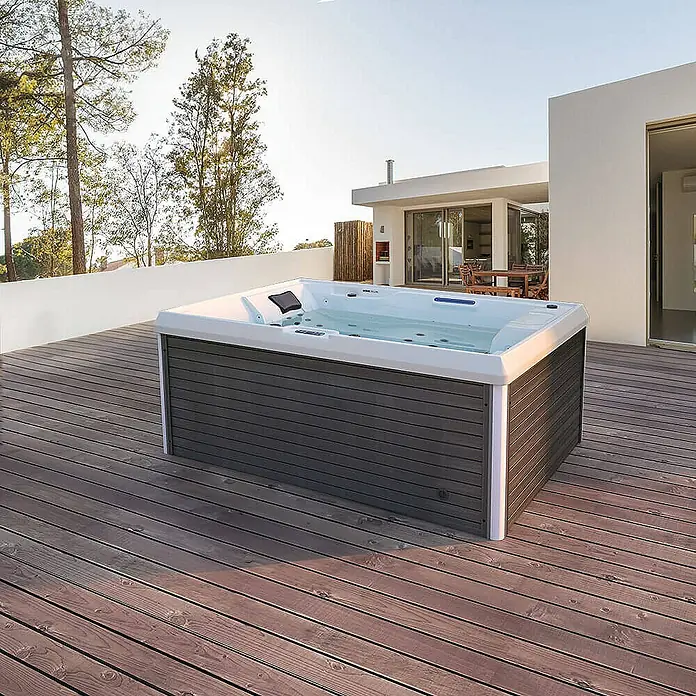 Home Deluxe Außen-Whirlpool STREAM PURE - Anthrazit, Max. Personenzahl: 3 Stk. - Ein luxuriöser Whirlpool mit grauer Verkleidung steht auf einer Holzterrasse, umgeben von Bäumen und einem modernen Haus.