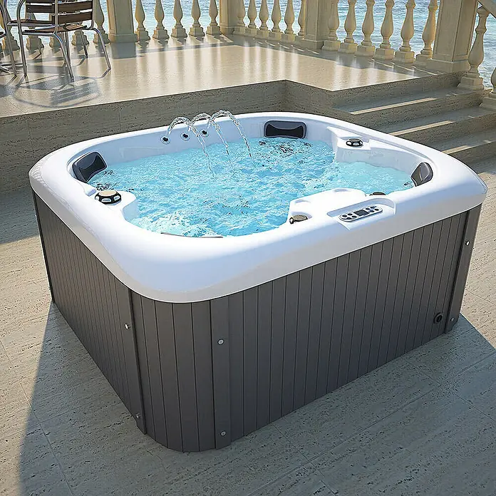 Home Deluxe Outdoor Whirlpool SEA STAR Mit Treppe und Thermoabdeckung -  - Ein luxuriöser Whirlpool mit Wasserspielen steht auf einer Terrasse mit Meerblick. Der Pool hat eine graue Verkleidung und ein weißes Inneres.