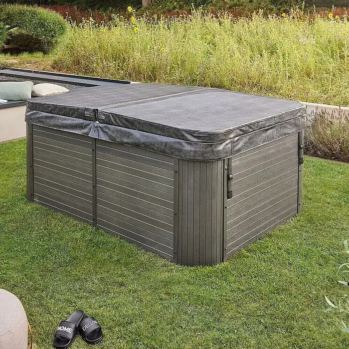 Home Deluxe Outdoor Whirlpool BEACH Mit Treppe und Thermoabdeckung -  - Ein abgedeckter Whirlpool im Garten, umgeben von Rasen und Pflanzen, lädt zum Entspannen ein. Die schwarzen Hausschuhe daneben.