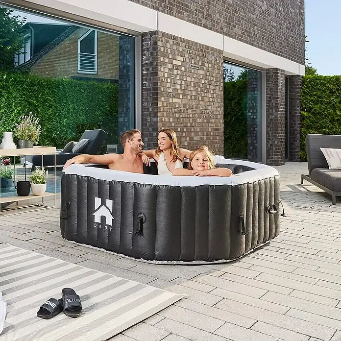 Home Deluxe Außen-Whirlpool DROP inkl. Abdeckung - 185 x 185 x 65 cm, Schwarz, Max. Personenzahl: 6 Stk. - Familie entspannt in einem aufblasbaren Whirlpool im Freien. Der Whirlpool ist schwarz mit weißem Rand und steht auf einer Terrasse.