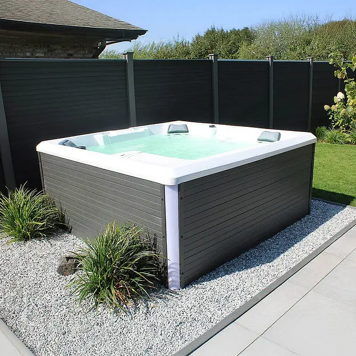 Home Deluxe Außen-Whirlpool STREAM BIG PURE - Anthrazit, Max. Personenzahl: 5 Stk. - Moderner Whirlpool im Garten mit grauem Gehäuse und weißem Rand, umgeben von Kies und Pflanzen.
