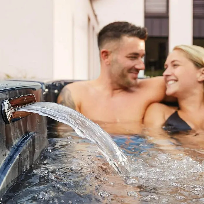 HOME DELUXE - Outdoor Whirlpool BLACK MARBLE - Mit Treppe und Thermoabdeckung | Ausstattung Mit Treppe und Thermoabdeckung -  - Whirlpool-Szene mit Wasserfall: Entspannung pur. Ein Paar genießt das luxuriöse Ambiente und die wohltuende Massage im sprudelnden Wasser.