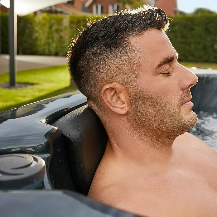 HOME DELUXE - Outdoor Whirlpool BLACK MARBLE - Mit Treppe und Thermoabdeckung | Ausstattung Mit Treppe und Thermoabdeckung -  - Ein Mann entspannt im Whirlpool mit Kopfstütze, die Komfort bietet und den Nacken stützt, während er die Ruhe genießt.