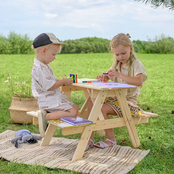 Timbela Kinder-Picknicktisch - 81 x 60 x 50 cm, Holz, Ohne Staufach - Kinder sitzen an einem Picknicktisch aus Holz und malen mit bunten Stiften auf Papier. Im Hintergrund ist eine grüne Wiese.