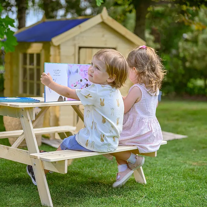 Timbela Kinder-Sitzgruppen-Set - 90 x 90 x 50 cm, Holz, Ohne Staufach - Kinder sitzen an einem hellen Holz-Picknicktisch mit einem blauen Buch und einem bunten Bilderbuch im Garten vor einem Spielhaus.