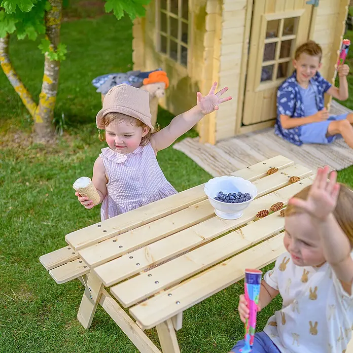 Timbela Kinder-Sitzgruppen-Set - 90 x 90 x 50 cm, Holz, Ohne Staufach - Eine helle Holz-Kindersitzgarnitur im Garten, umgeben von spielenden Kindern, die Eis und Blaubeeren genießen.