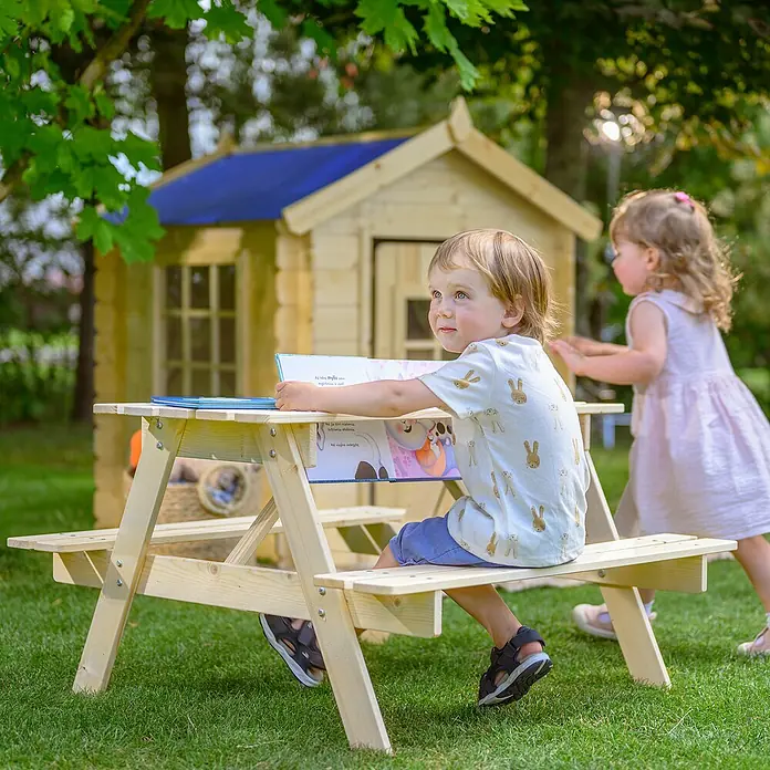 Timbela Kinder-Sitzgruppen-Set - 90 x 90 x 50 cm, Holz, Ohne Staufach - Kind sitzt am Picknicktisch für Kinder, ein Buch aufgeschlagen, mit einem Spielhaus im Hintergrund, ideal für Outdoor-Spaß.