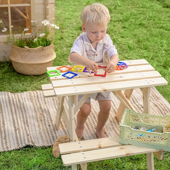 Timbela Kinder-Picknicktisch - 81 x 60 x 50 cm, Holz, Ohne Staufach - Ein Kind spielt am Holztisch mit bunten Spielzeugquadraten im Garten, während eine Pflanze und eine Spielzeugkiste die Szene ergänzen.