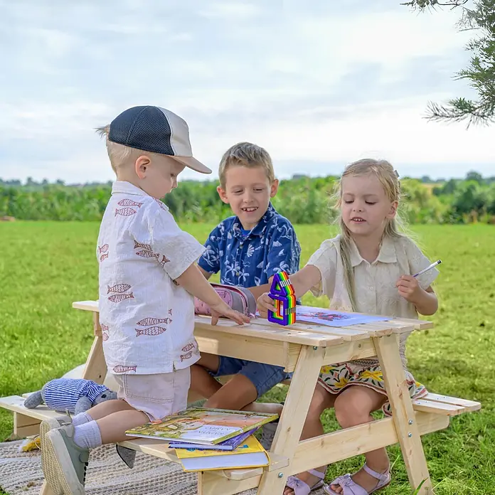 Timbela Kinder-Sitzgruppen-Set - 90 x 90 x 50 cm, Holz, Mit Staufach - Drei Kinder sitzen an einem Picknicktisch aus Holz im Freien, wo sie spielen und Bücher lesen.
