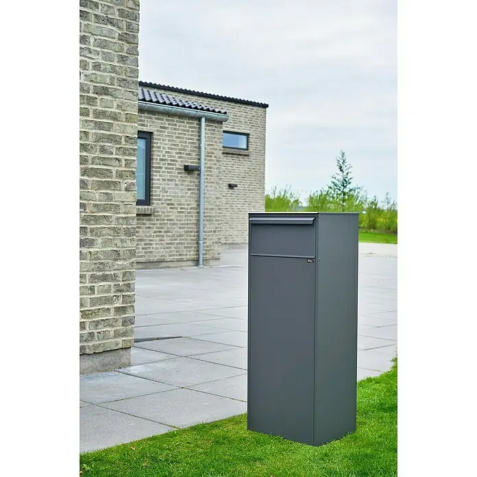 Allux Paketbox 800 - L x B x H: 280 x 390 x 980 mm, Anthrazit, Stahl, Paketfach, Entnahme: Hinten - Ein moderner, anthrazitfarbener Briefkasten steht auf dem Rasen vor einem Backsteinhaus und bietet eine stilvolle Möglichkeit, Post zu empfangen.