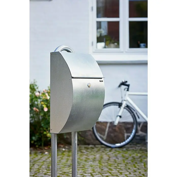 Allux Briefkasten 7000 - L x B x H: 195 x 320 x 510 mm, Silber, Stahl - Nahaufnahme eines modernen, silberfarbenen Briefkastens auf zwei Ständern vor einem weißen Haus mit einem Fahrrad im Hintergrund.