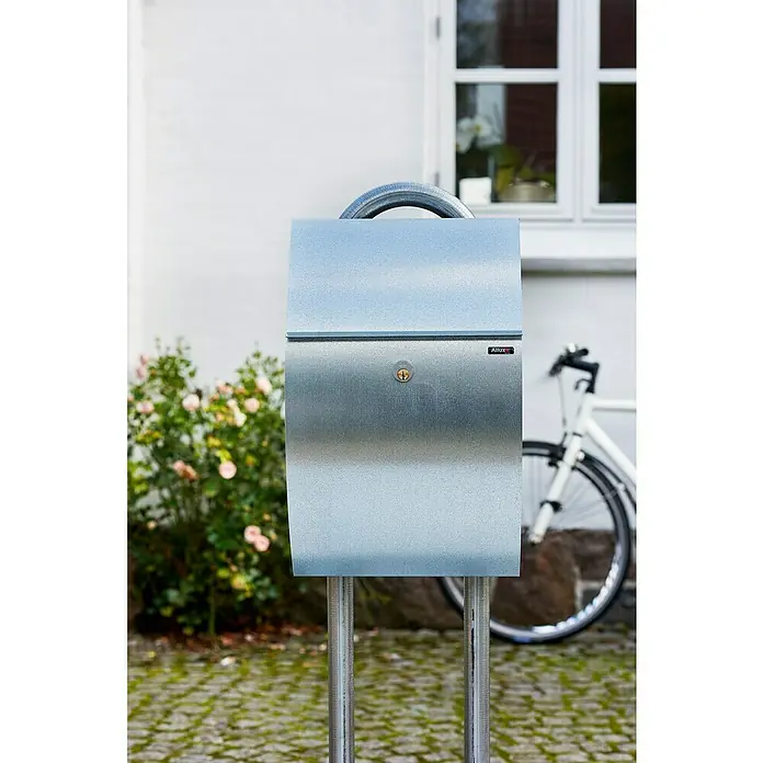 Allux Briefkasten 7000 - L x B x H: 195 x 320 x 510 mm, Silber, Stahl - Nahaufnahme eines modernen Allux Briefkastens aus Edelstahl mit Schloss und Aufhängevorrichtung vor einem Haus.