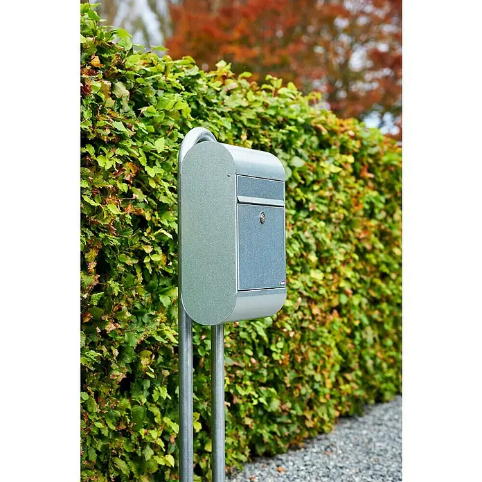 Allux Standbriefkasten 6000 - L x B x H: 170 x 345 x 485 mm, Silber, Stahl - Nahaufnahme eines modernen silbernen Briefkastens mit Briefschlitz und Schloss, montiert auf zwei Pfosten vor einer grünen Hecke.