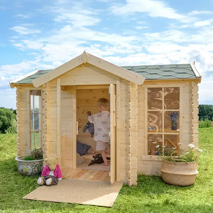 Timbela Spielhaus - L x B x H: 187 x 241 x 151 cm, Holz, Natur - Ein helles Holzspielhaus mit grünen Dachschindeln, das von einem Kind im Pyjama betreten wird, umgeben von Grün und Blumentöpfen.
