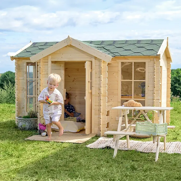 Timbela Spielhaus - L x B x H: 187 x 241 x 151 cm, Holz, Natur - Ein hölzernes Kinderspielhaus im Garten mit einem Jungen davor, der Spielzeug hält, und einem Tisch mit Bank.